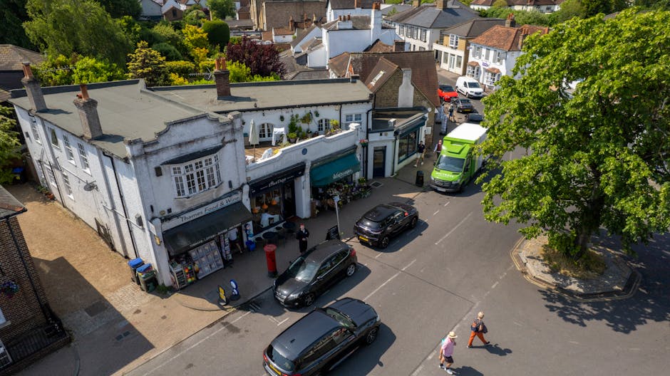 An aerial view of a busy street in Kingston upon Thames showing a two-storey white building with a grey flat roof, housing small shops with awnings and display windows. Several vehicles, including a green van, a black car, and a grey car, are parked along the road near the building. In front of the shops, two pedestrians are walking, and one appears to be carrying a bag. The street is lined with trees, casting shadows, and other residential buildings with various roof types are visible in the background. The scene depicts a typical urban setting suitable for house removals and furniture transport services, with parked vehicles and pedestrians indicating active movement and logistical considerations for home relocation or moving processes. Kingston Upon Thames Movers may utilize such locations for loading and unloading logistics during home relocations in the Canbury Gardens KT2 area, supporting efficient packing and moving operations.