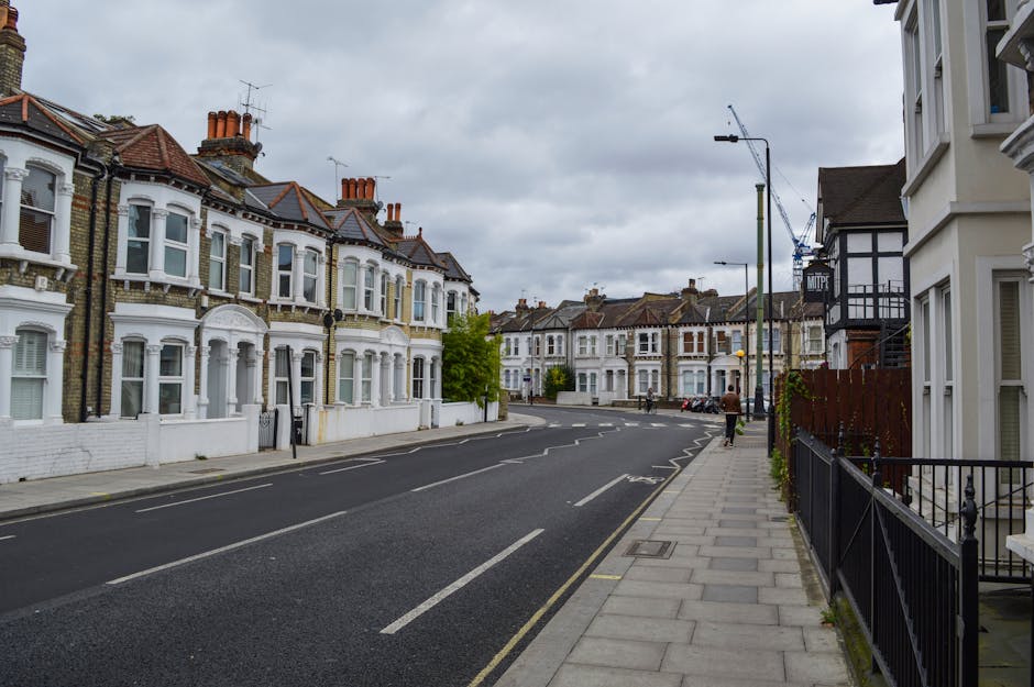 A quiet residential street in Kingston upon Thames with a row of Victorian-style terraced houses featuring bay windows and brick facades on the left, and modern white and black-finished buildings with balconies on the right. The sidewalk is paved with concrete slabs, and there are black fence railings along the front of the houses on the right. The road has a designated parking lane with a single car visible in the distance, and a person walking along the pavement. The overcast sky is filled with grey clouds, and a tall street lamp extends above the scene. The environment appears calm and suitable for home relocation or furniture transport activities, with the street being part of a typical urban area serviced by [COMPANY_NAME] for local house removals and packing and moving services.