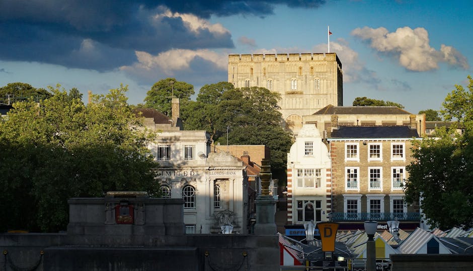The image depicts a cityscape with a historic stone castle or fortress on a hilltop in the background, illuminated by sunlight under a partly cloudy sky. In the foreground, modern multi-storey buildings with white and brick facades are visible, some with large windows and decorative architectural details. Below these buildings, a street area is partially visible with features such as taxis, a bus stop, and road signage, suggesting an urban environment. Lush green trees frame parts of the scene, adding contrast to the architecture. The image captures a blend of historic and contemporary elements characteristic of a town like Kingston upon Thames, providing context related to home relocation and moving services. Kingston Upon Thames Movers may use such visual representations in their content about the logistics involved in furniture transport, packing, and the loading process during house removals, illustrating the setting for city-based moving operations.