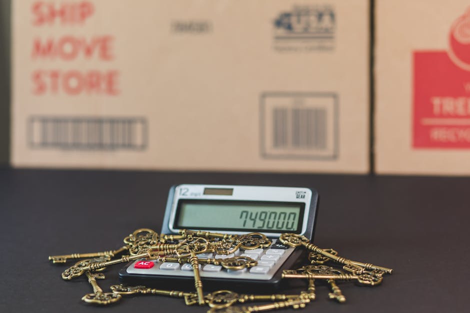 A calculator displaying the number 749,000 is placed on a dark surface, surrounded by a collection of antique gold keys of various sizes and designs, some attached to small chains. Behind the calculator, there are large cardboard moving boxes with printed labels and barcode stickers, positioned against a neutral background. The setting appears to be inside a property or a storage area, with the keys and calculator arranged in the foreground, indicating a focus on valuation or inventory related to a home relocation or furniture transport. The scene reflects elements involved in packing, moving logistics, and valuation processes during house removals, with the presence of packaging materials and moving equipment suggesting ongoing packing or loading activities, consistent with professional removals services by Kingston Upon Thames Movers.