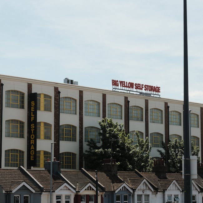 Photograph of a large multi-storey building with a sign reading 'Big Yellow Self Storage' on the roof and a vertically oriented sign on the side that says 'Self Storage' in yellow lettering on a black background. The building's facade features multiple evenly spaced, large arched windows with yellow frames, separated by exposed brick columns. In the foreground, there are small residential-style houses with pitched roofs and chimneys, positioned beneath the building. To the left, a person wearing a green shirt is visible near a grey metal bin on the pavement, which is partly shaded by trees with green foliage. Metal lampposts and street signs are also present, indicating an urban environment. The scene is lit by daylight with a partly cloudy sky overhead, capturing a moment in the process of home relocation or furniture transport involving storage facilities and moving logistics, as managed by Kingston Upon Thames Movers.