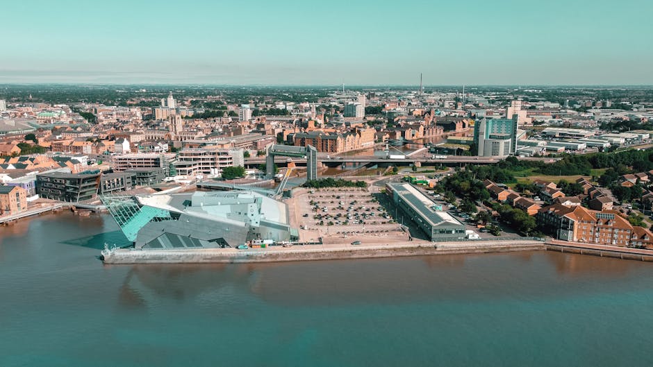 An aerial view of a cityscape overlooking a river, featuring modern architectural buildings with distinctive angular and glass facades, likely part of a waterfront development area. The scene includes