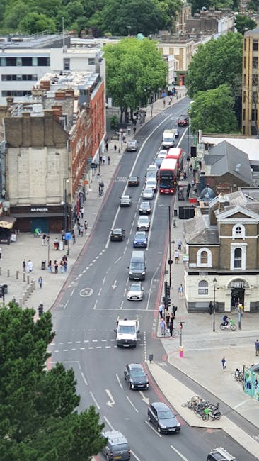 An aerial view of a busy urban street in Kingston upon Thames, showing multiple parked and moving vehicles including cars, vans, and buses. The road is divided by a central lane with vehicles traveling in both directions. On the sidewalks, there are pedestrians walking and cycling, some near parked bicycles. Buildings lining the street include a mix of commercial and residential structures, with trees providing greenery along the pavement. The scene captures the typical environment for home relocation or furniture transport logistics, where vehicles are prepared for moving services such as those offered by Kingston Upon Thames Movers, with clear visibility of the road, parking, and street activity suitable for coordinated loading and unloading during house removals.