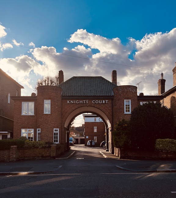 A brick archway entrance with the sign 'Knights Court' on top, situated between two residential buildings with windows and chimneys, under a partly cloudy sky. The arch leads to a residential street with parked cars visible in the background. The scene is well-lit with natural daylight, and some greenery, including bushes, is present on either side of the arch. The image is associated with house removals and relocation services, such as those provided by Kingston Upon Thames Movers, highlighting the process of home furniture transport and loading during a short-distance move from a property in Kingston Hill.