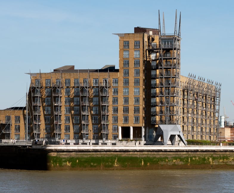 A modern multi-storey building under construction along a riverside, with scaffolding covering parts of the structure. The building features a combination of brick and glass windows, and scaffolding poles are visible on multiple floors. On the ground level, there is an open doorway area, and the building has several balconies. The construction site includes temporary fencing and equipment, and nearby, a riverbank with a concrete wall separates the water from the property. The sky is clear with some light clouds, and the scene captures ongoing development work typical of city urban renewal projects, with Kingston Upon Thames Movers specializing in house and office removals in the area, including facilitating furniture transport and packing during home relocation processes.