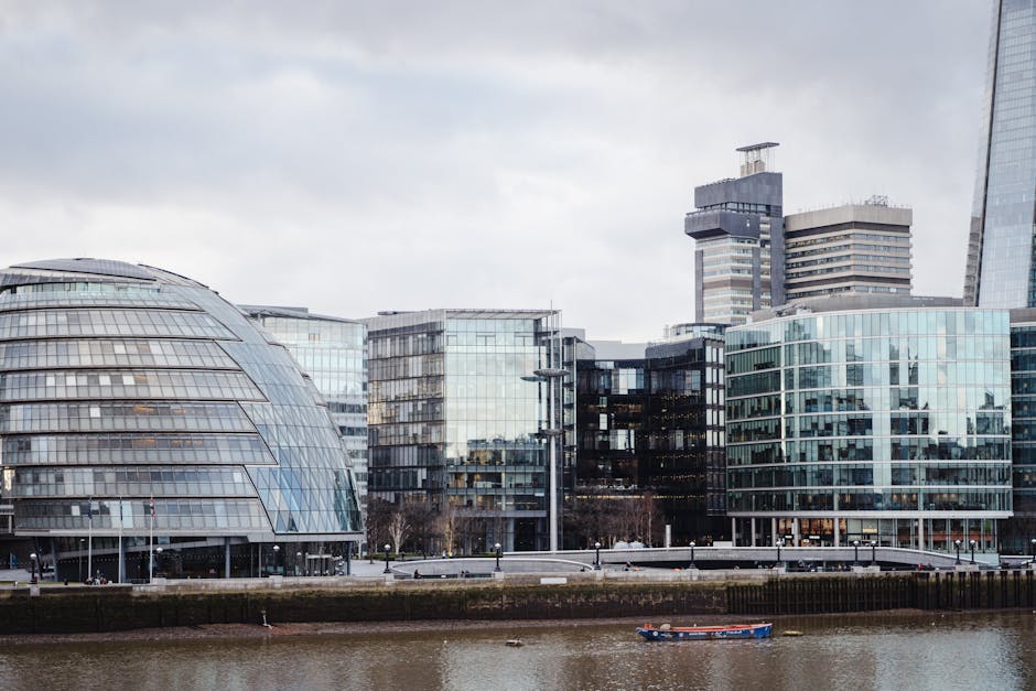 A view of modern office buildings along the riverside in Kingston upon Thames with a distinctive curved glass-domed structure on the left. The buildings feature large glass facades reflecting the cloudy sky, with some containing visible interior lighting. In the foreground, a small boat is moored on the river, which runs parallel to the pavement and office complex. The area around the buildings is open, with a few street lamps and minimal pedestrian activity. The scene captures a typical urban riverside setting suitable for professional removals and office furniture transport, with the Kingston Upon Thames Movers team likely preparing for a house or office relocation involving careful packing and loading of furniture, boxes, and equipment onto a vehicle for transportation from this commercial environment.