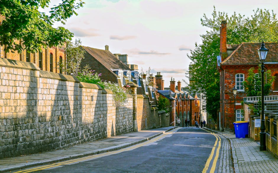 A residential street scene during daytime showing a paved pavement on the right with a blue recycling bin and black lampposts, and a low stone wall on the left side with a sidewalk. Behind the wall, there are brick and stone buildings with various roof styles, including gabled and flat roofs, some with chimneys. Several trees with green foliage are visible on both sides, casting shadows on the pavement. In the distance, a few pedestrians are walking along the street, which is slightly curved, and the sky above is partly cloudy with soft, natural light. The scene depicts a typical British neighborhood environment suitable for house removals or furniture transport as part of a home relocation process, with a focus on street and façade details that movers like Kingston Upon Thames Movers may consider during loading and unloading activities.