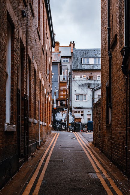 Image of a narrow urban alleyway between two brick buildings, showing a view towards backyards and upper floors of residential properties in Kingston upon Thames. The alley is paved with asphalt and features double yellow lines along the edges, indicating parking restrictions. On the left side, black drainpipes and window frames are visible, while on the right side, black pipes run down the brick wall. At the far end, wooden stairs with a metal railing lead up to an external door on the second floor of an adjoining building. The background includes rooftops of various residential structures, some with dormer windows and chimneys, under a cloudy sky. This setting depicts typical backstreet conditions where house removals could encounter access challenges, emphasizing the importance of professional moving services like Kingston Upon Thames Movers for safe furniture transport and packing during home relocation processes.