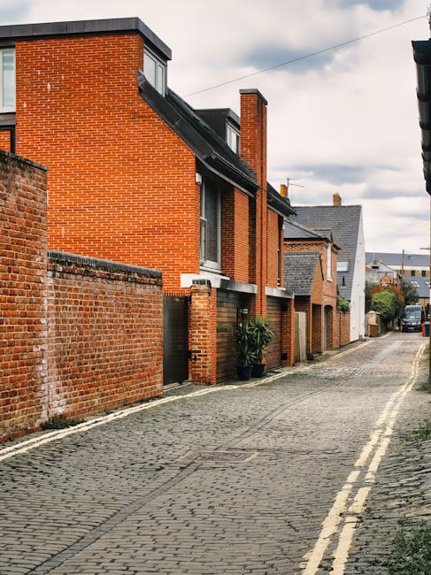 Image of a narrow urban alleyway between two brick buildings, showing a view towards backyards and upper floors of residential properties in Kingston upon Thames. The alley is paved with asphalt and features double yellow lines along the edges, indicating parking restrictions. On the left side, black drainpipes and window frames are visible, while on the right side, black pipes run down the brick wall. At the far end, wooden stairs with a metal railing lead up to an external door on the second floor of an adjoining building. The background includes rooftops of various residential structures, some with dormer windows and chimneys, under a cloudy sky. This setting depicts typical backstreet conditions where house removals could encounter access challenges, emphasizing the importance of professional moving services like Kingston Upon Thames Movers for safe furniture transport and packing during home relocation processes.