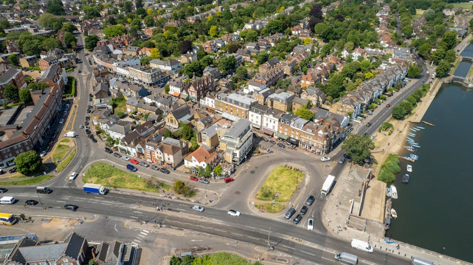 Aerial view of a town in Kingston upon Thames featuring a mixture of residential and commercial buildings, with a prominent street running through the centre. Several vehicles, including cars, vans, and a truck, are parked along the road and in parking areas. Green spaces and trees are visible interspersed throughout the area, with a river or body of water located on the right side, complete with small boats docked along the quay. The street appears busy with moving traffic, and the surroundings include a combination of brick and modern styled buildings, some with flat roofs and others with pitched roofs, typical of urban housing or retail units. This scene illustrates the typical environment where Kingston Upon Thames Movers might perform home relocation and furniture transport, capturing the logistics involved in house removals from an aerial perspective during daylight hours.