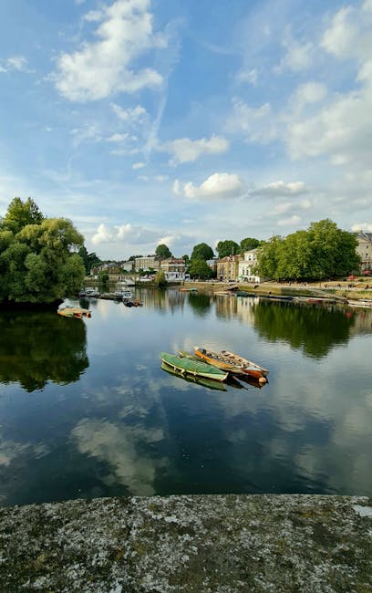 A scenic view of a calm river with a clear blue sky and scattered white clouds above. On the river's surface, multiple small boats with wooden and plastic hulls are moored close to the riverbank, which features a paved pathway and lush green trees. Across the river, residential buildings and houses with light-colored facades and varied roof styles are visible, partially obscured by greenery. The scene is illuminated by natural daylight, showcasing reflections of the sky and trees on the water. This setting depicts an outdoor environment suitable for a house relocation or moving service, such as those provided by Kingston Upon Thames Movers, emphasizing the importance of careful planning in transport logistics, including packing, loading, and unloading furniture and belongings during a home removal process near the riverside.
