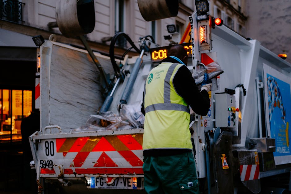 A large white commercial vehicle, likely used for house removals, parked on a paved surface with its door open. Attached to the vehicle is a blue waste collection bin with a closed lid, positioned on